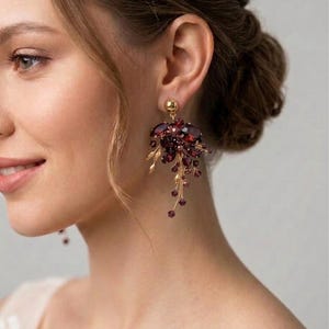May include: A gold-toned statement earring featuring a cluster of deep red, faceted crystals and small, dangling dark red beads. The design includes delicate gold-colored metal branches and leaves, with a smooth gold sphere at the earlobe attachment.