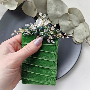 May include: A decorative hair accessory featuring a gold-toned wire frame adorned with clear teardrop crystals, dark green beads, and small sparkling accents. The piece is attached to a rectangular base wrapped in vibrant green velvet ribbon.