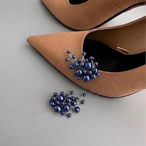 May include: A pair of tan pointed-toe heels with a cluster of dark blue pearl embellishments. One shoe has the embellishment attached, and the other shoe and a separate embellishment are visible. The shoes have a classic, elegant design.