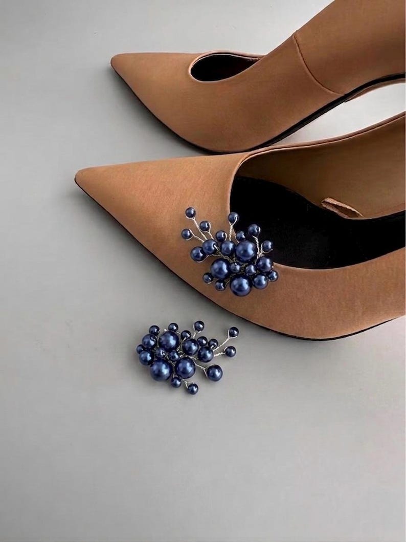May include: A pair of tan pointed-toe heels with a cluster of dark blue pearl embellishments. One shoe has the embellishment attached, and the other shoe and a separate embellishment are visible. The shoes have a classic, elegant design.