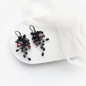 May include: A pair of dangle earrings featuring clusters of small black beads and faceted dark purple teardrop crystals. The components are arranged on thin black wire stems, creating a cascading, branch-like structure with hook-style ear wires.
