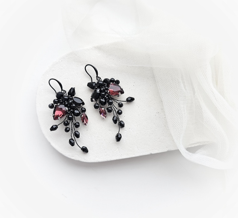 May include: A pair of dangle earrings featuring clusters of small black beads and faceted dark purple teardrop crystals. The components are arranged on thin black wire stems, creating a cascading, branch-like structure with hook-style ear wires.