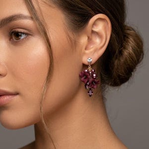 May include: A dangling earring featuring a small clear crystal stud at the top, connected to a cluster of deep burgundy floral-shaped elements, round dark purple pearls, and a single faceted marquise-cut crystal drop at the bottom.