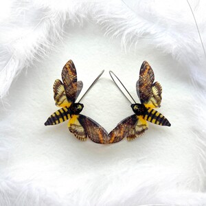 May include: A pair of dangle earrings featuring a detailed, realistic depiction of a Death's-head Hawkmoth, a black and yellow moth with a skull-like pattern on its thorax. The earrings are made of metal and have a hook closure.