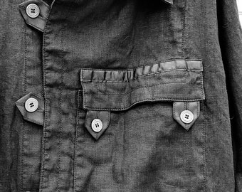 Puede incluir: Una chaqueta de lino negra con un bolsillo grande en la parte delantera, un cinturón y varios botones. La chaqueta tiene cuello y mangas largas.