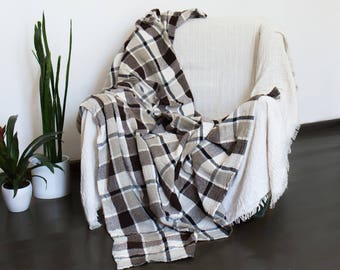 May include: A plaid blanket in shades of brown, gray, and white draped over a white armchair. The blanket has a classic check pattern. The chair is set against a white wall with potted plants in the background. The blanket appears soft and inviting.