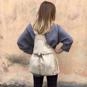 Puede incluir: Una mochila de lona beige con cierre de cordón y correas ajustables. La mochila la lleva puesta una persona con el pelo largo y castaño, de pie frente a una pared texturizada.