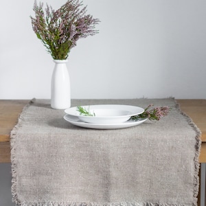 May include: A light brown linen table runner with frayed edges. A white plate with a sprig of rosemary sits on top of the runner. A white vase with a bouquet of pink heather sits on a wooden table.