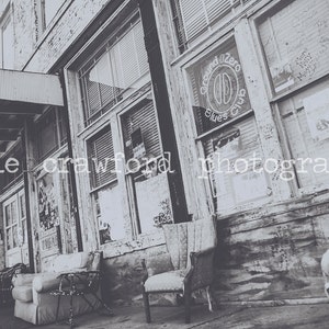 May include: A black and white photo of a building with a sign that reads "Ground Zero Blues Club". The building has large windows and a wooden awning. There are two chairs in the foreground.