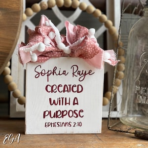 May include: White wooden block with the text "Sophia Raye Created With A Purpose Ephesians 2:10" in maroon lettering. The block is decorated with a pink and white fabric with white pom-poms.