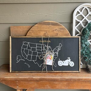 May include: A black chalkboard with a white outline of the United States map. The map is labeled "J.A.F.F.J.A." and has a white silhouette of a motorcycle on the right side. The chalkboard is framed in wood and is sitting on a wooden table.