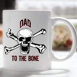 May include: White ceramic mug with a black and white skull and crossbones design. The text "DAD TO THE BONE" is printed in red below the skull.