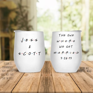 May include: Two white stainless steel wine tumblers with black text. The tumbler on the left says "JESS & SCOTT" with dots between the letters. The tumbler on the right says "THE ONE WHERE WE GET MARRIED 9-28-19".