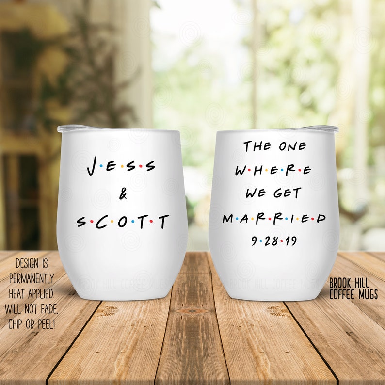 May include: Two white stainless steel wine tumblers with black text. The tumbler on the left says "JESS & SCOTT" with dots between the letters. The tumbler on the right says "THE ONE WHERE WE GET MARRIED 9-28-19".