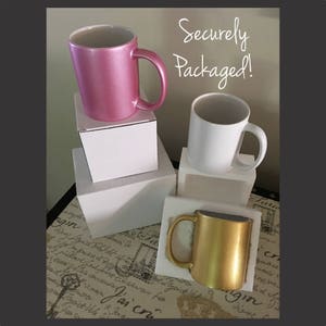 May include: Three ceramic mugs in pink, white, and gold are displayed on white boxes. The text "Securely Packaged!" is written in black marker on a white background.