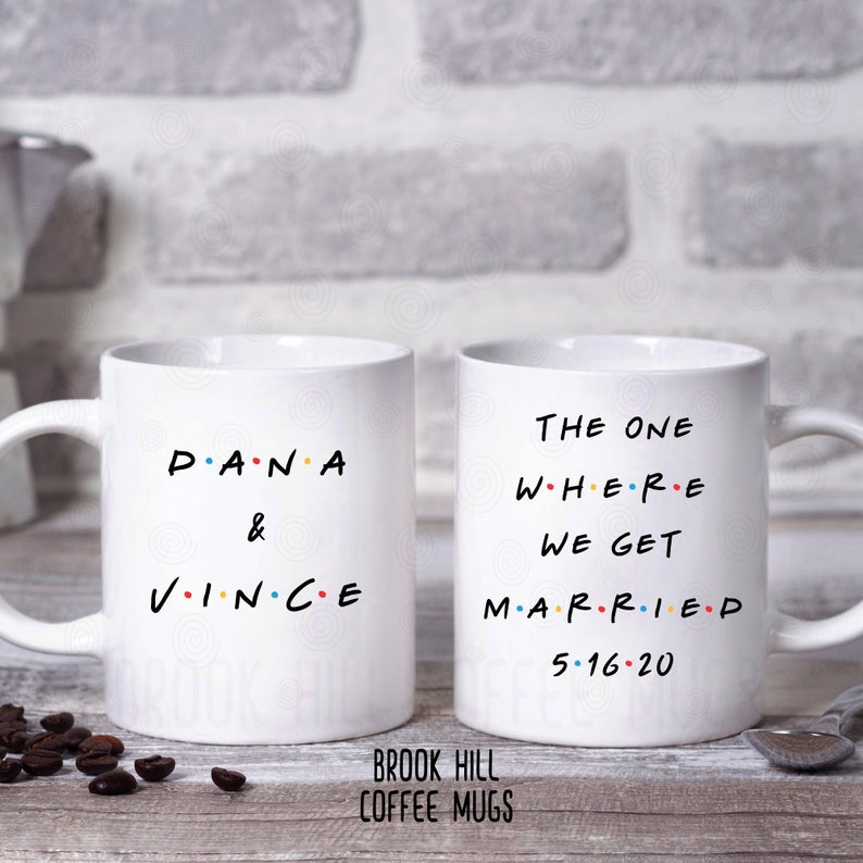 May include: Two white coffee mugs with black text. The mug on the left says "P.A.N.A & V.I.N.C.E". The mug on the right says "THE ONE WHERE WE GET MARRIED 5.16.20".