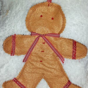 Może przedstawiać: Choinkowa ozdoba w kształcie piernikowego ludzika wykonana z brązowego filcu z czerwonymi i białymi kratkowanymi wstążkami. Ozdoba ma pętelkę na górze do zawieszenia.