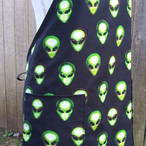 May include: Black apron with a repeating pattern of green alien faces. The apron has a large pocket and adjustable neck strap. The alien faces have large black eyes and are outlined in green.
