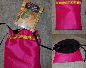 Puede incluir: Una bolsa de cordón de satén rosa con forro negro y ribete dorado. La bolsa está vacía y lista para guardar objetos. La bolsa está sobre una superficie gris texturizada.