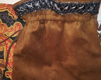 May include: A brown suede drawstring pouch with a decorative silver and black trim around the opening. The pouch is partially open, revealing a colorful paisley pattern fabric lining.
