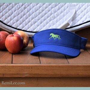 May include: A royal blue visor with a green horse embroidered on the front. The visor is lying on a wooden surface with red apples in the background.