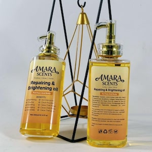 May include: Two bottles of AMARA SCENTS Repairing & Brightening Oil for face and body. The bottles are clear with gold pumps and labels. The labels have text and are filled with a yellow liquid. The bottles are in front of a black and gold geometric candle holder. Each bottle contains 10.14 fl oz.
