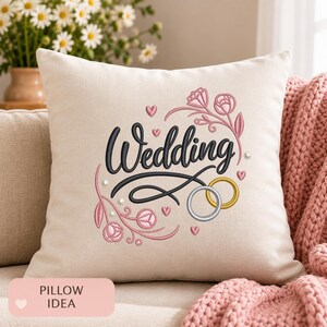 Puede incluir: Un cojín cuadrado de color blanco roto con el texto Wedding bordado en negro. El diseño incluye enredaderas florales bordadas en rosa, corazones pequeños y dos anillos entrelazados, uno plateado y otro dorado, con pequeñas perlas blancas.