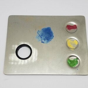 May include: A metal paint palette with four circular wells. The wells are filled with blue, red, yellow, and green paint.