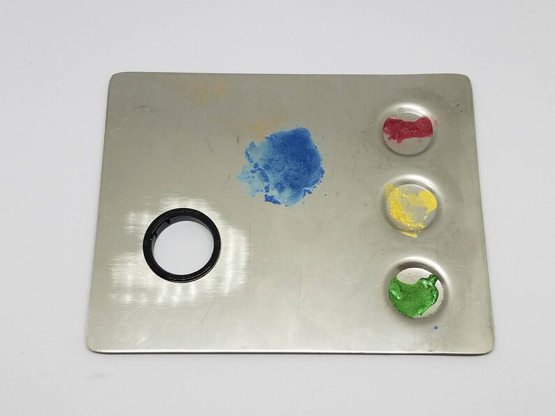 May include: A metal paint palette with four circular wells. The wells are filled with blue, red, yellow, and green paint.