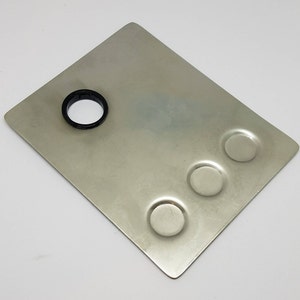 May include: A silver metal palette with three round wells and a larger circular hole in the center. The palette is designed for mixing paints or other materials.