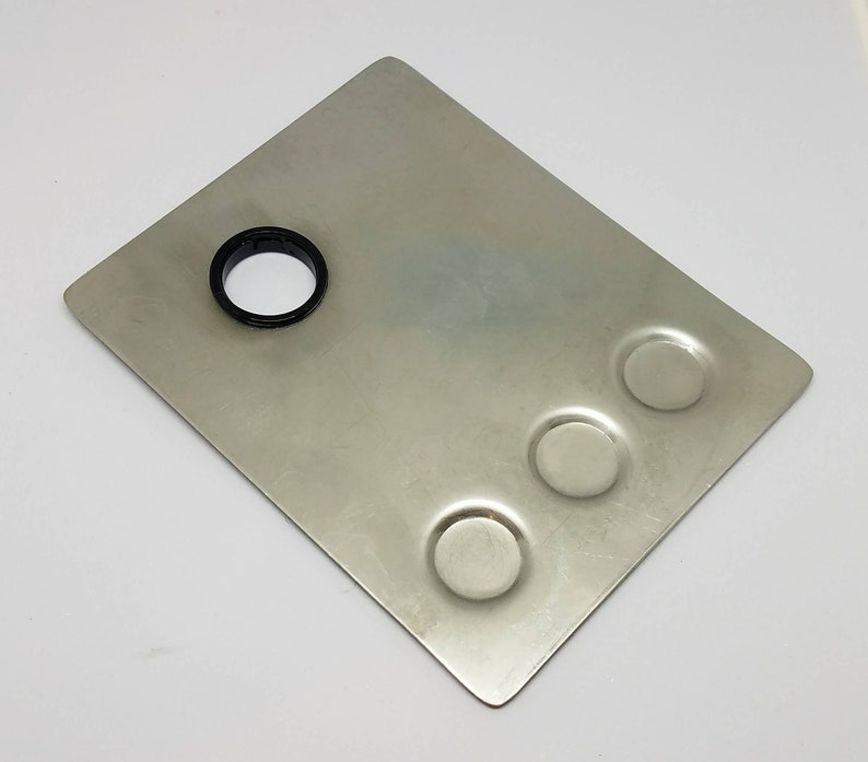 May include: A silver metal palette with three round wells and a larger circular hole in the center. The palette is designed for mixing paints or other materials.