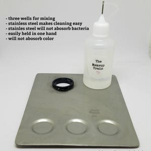 May include: A stainless steel mixing palette with three wells for mixing makeup. The palette is on a silver surface with a clear bottle of mixing medium labeled "The Makeup Train".