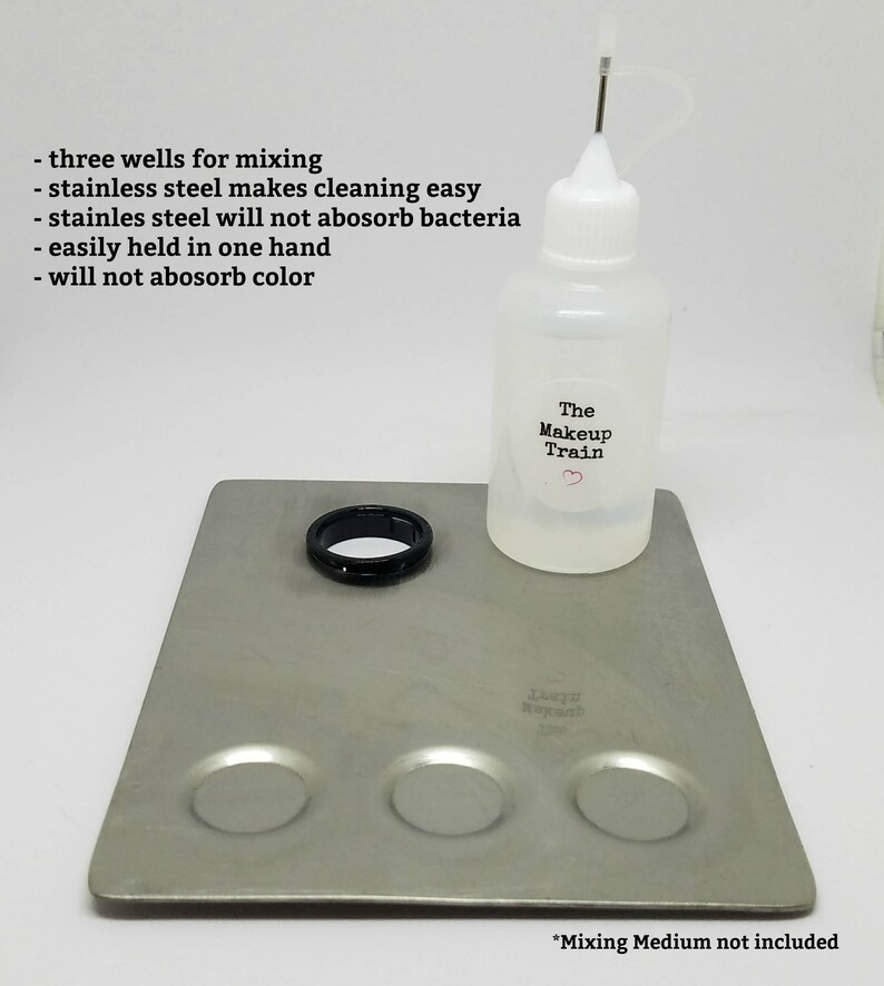 May include: A stainless steel mixing palette with three wells for mixing makeup. The palette is on a silver surface with a clear bottle of mixing medium labeled "The Makeup Train".