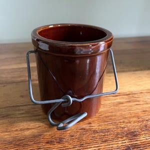May include: A small, brown ceramic crock with a shiny glaze and a wire handle. The crock is cylindrical with a slightly flared rim. The wire handle is attached to the sides of the crock. The background is a wooden surface.
