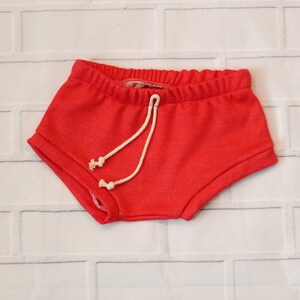 May include: Red shorts with a white drawstring. The shorts are made of a soft, textured fabric.
