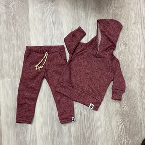 May include: A burgundy two-piece tracksuit set. The set includes a hooded sweatshirt with a zip and matching tracksuit bottoms with a drawstring. The fabric has a speckled texture. The set is designed for children.