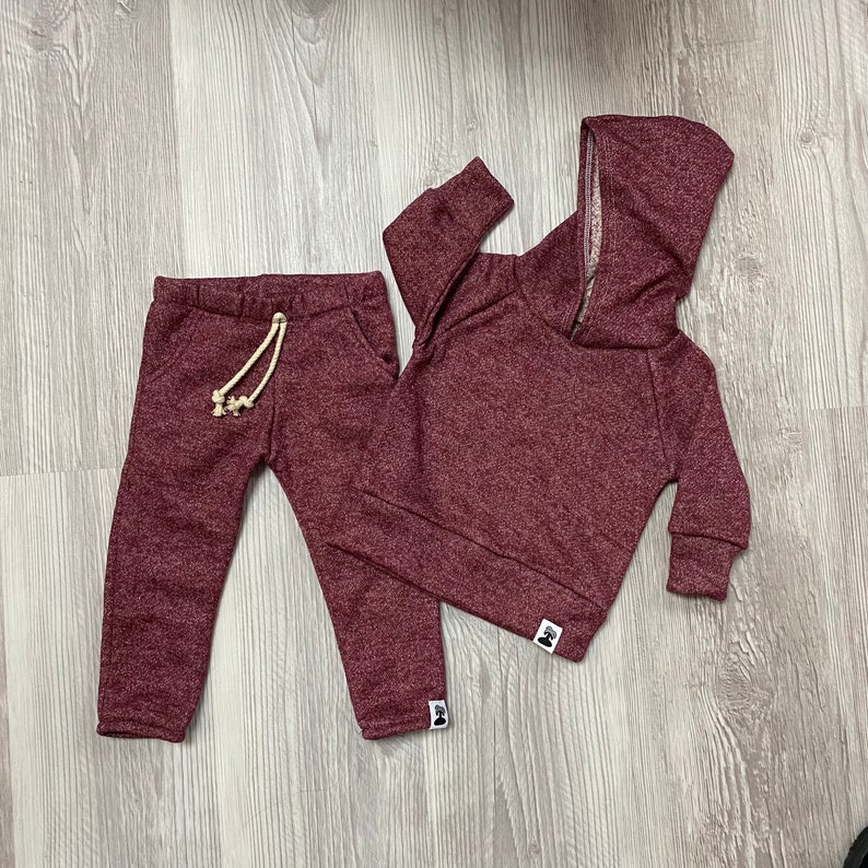 May include: A burgundy two-piece tracksuit set. The set includes a hooded sweatshirt with a zip and matching tracksuit bottoms with a drawstring. The fabric has a speckled texture. The set is designed for children.