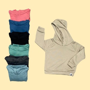 May include: A collection of children's hoodies in various colors, including pink, blue, black, and green, are stacked on the left. A single tan hoodie is on the right, showcasing the design. The hoodies have a simple, casual style with a hood and long sleeves.