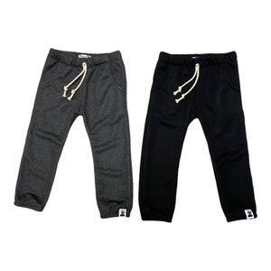 May include: Two pairs of children's sweatpants. One pair is dark gray, the other is black. Both have an elastic waistband with a cream-colored drawstring, front pockets, and a small white tag on the left leg cuff.