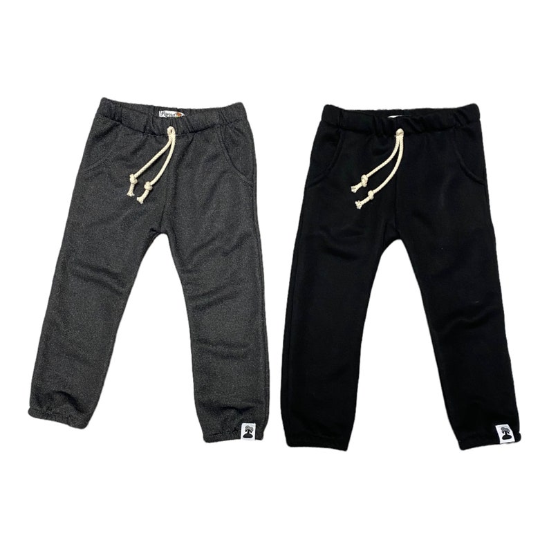 May include: Two pairs of children's sweatpants. One pair is dark gray, the other is black. Both have an elastic waistband with a cream-colored drawstring, front pockets, and a small white tag on the left leg cuff.