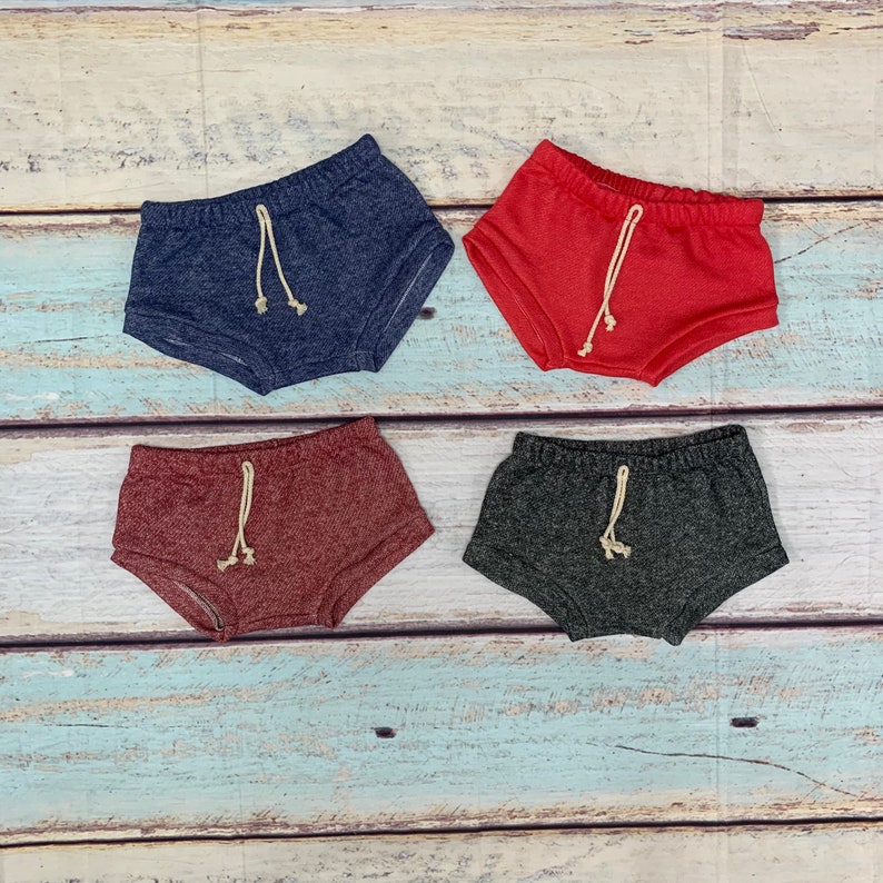 May include: Four pairs of toddler shorts in different colors. The shorts are made of a soft, stretchy fabric and have a drawstring waistband. The colors are navy blue, red, burgundy, and gray.