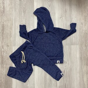 May include: A navy blue children's tracksuit set, comprising a hooded sweatshirt and matching tracksuit bottoms. The set has a speckled texture and a drawstring on the trousers. The sweatshirt and trousers have a small logo at the bottom.