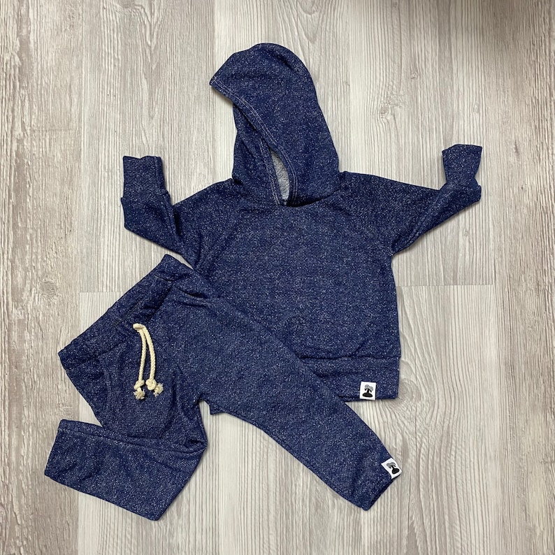 May include: A navy blue children's tracksuit set, comprising a hooded sweatshirt and matching tracksuit bottoms. The set has a speckled texture and a drawstring on the trousers. The sweatshirt and trousers have a small logo at the bottom.
