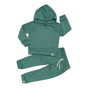 May include: A two-piece children's outfit in a muted teal color. The set includes a hooded sweatshirt with long sleeves and matching sweatpants. The pants have a drawstring waist and the set has small white tags.