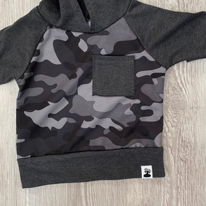 May include: A gray and black camouflage hoodie with a gray pocket and a small gray tag on the bottom.
