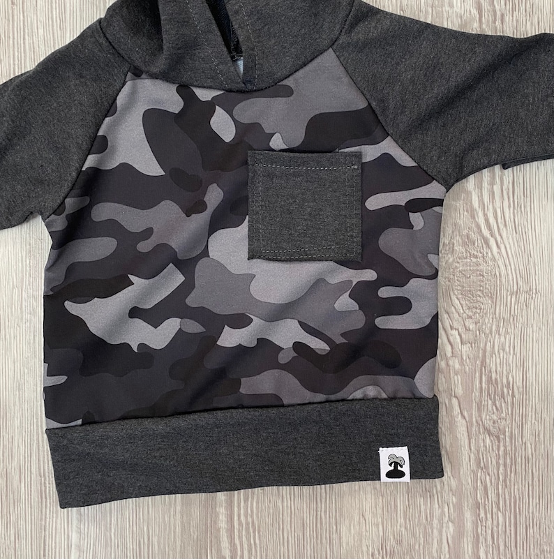 May include: A gray and black camouflage hoodie with a gray pocket and a small gray tag on the bottom.