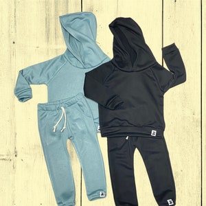 May include: Two matching children's outfits. One is a light blue hoodie and sweatpants set with a drawstring waist. The other is a black hoodie and sweatpants set. Both sets have a small white tag with a logo.