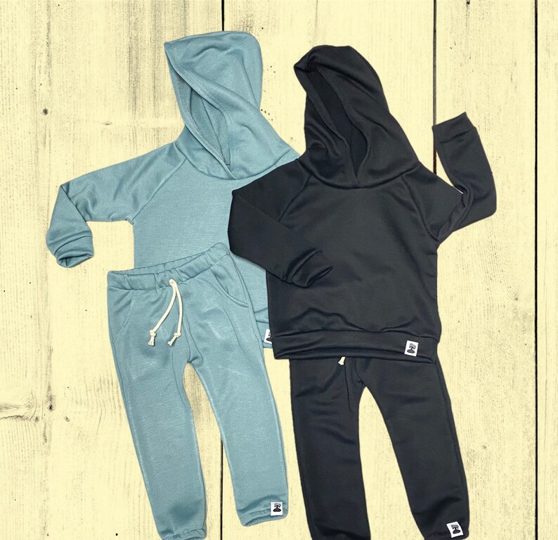 May include: Two matching children's outfits. One is a light blue hoodie and sweatpants set with a drawstring waist. The other is a black hoodie and sweatpants set. Both sets have a small white tag with a logo.