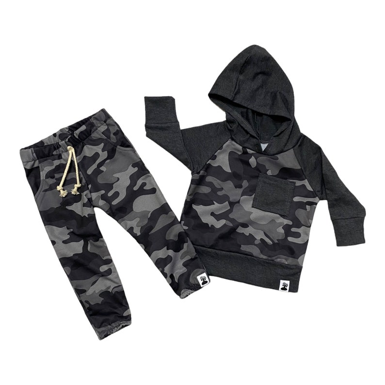 May include: A black and gray camouflage hoodie and matching sweatpants set for a baby or toddler. The hoodie has a drawstring hood and a small pocket on the front. The sweatpants have a drawstring waistband.