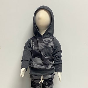 May include: A gray hooded sweatshirt with a camouflage pattern on the front. The sweatshirt has a pocket on the front and a drawstring hood.